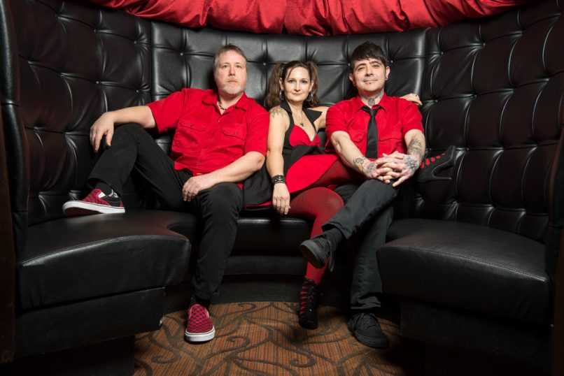 Band in matching red clothing seated in front of dark alt background